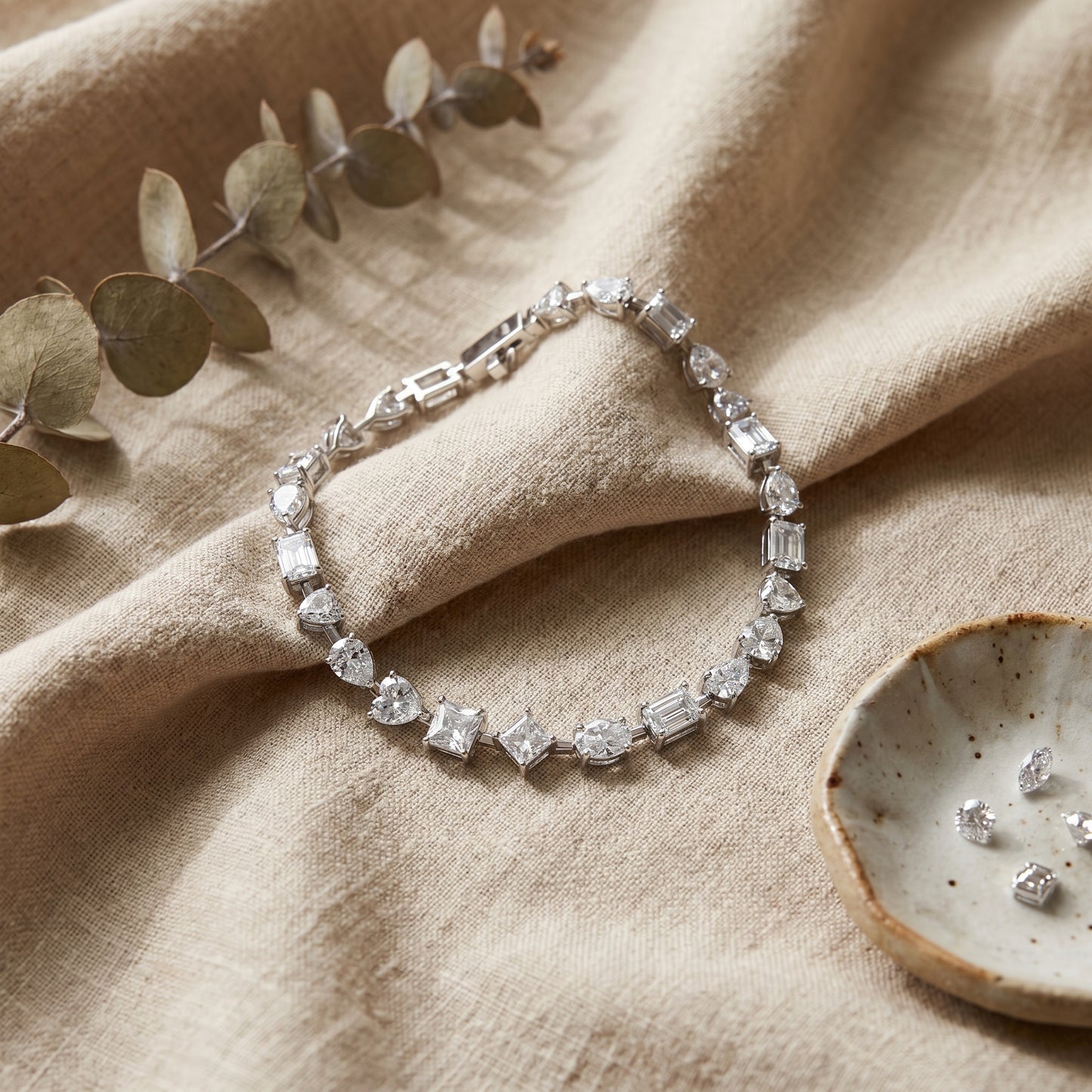 A shimmering multi-shape moissanite eternity bracelet beautifully laid on a neutral-toned textured linen background.