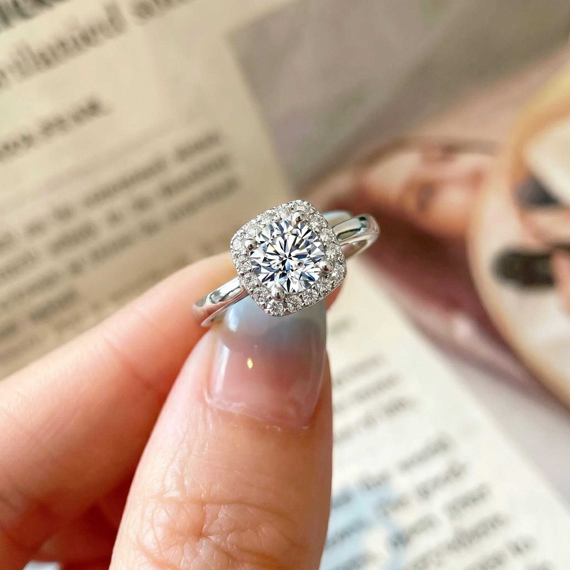 Hand holding cushion halo moissanite ring near a book in natural light